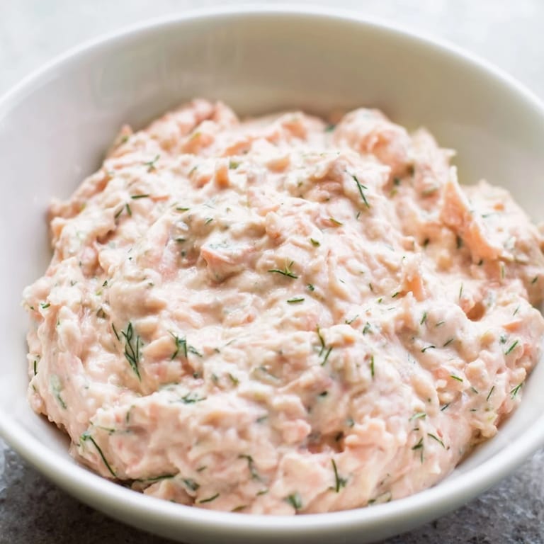 Smooth, pink smoked salmon dip spread on a crispy bagel chip, topped with a sprig of dill for a perfect appetizer bite.